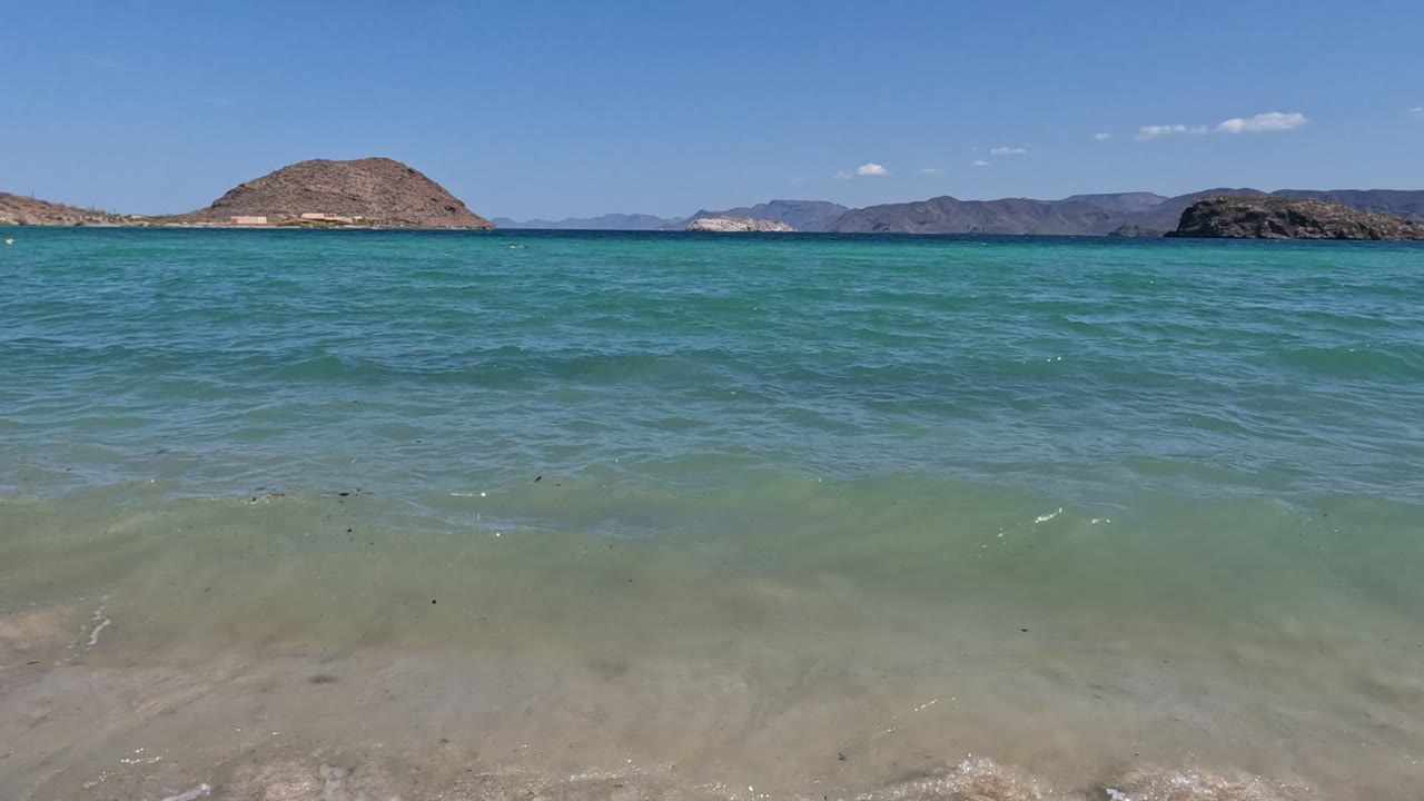 At 0:00 in Baja California, Mexico, a low shoreline view shows turquoise water, pale sand, distant rocky hills, scattered clouds, and no visible people.