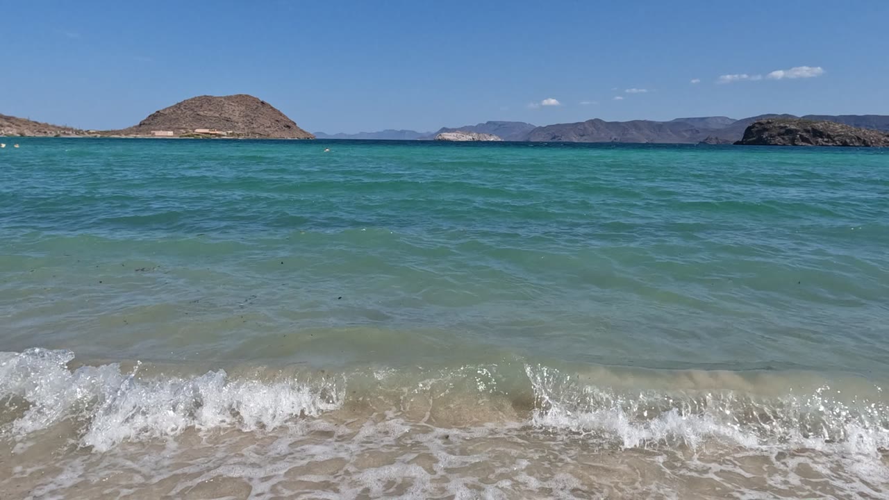 Foamy wave spreads across pale sand with turquoise sea and low islands on the horizon.