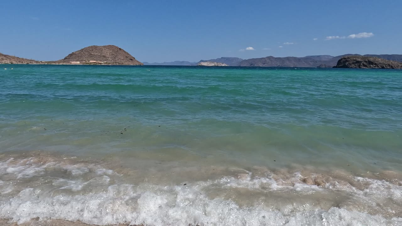 At 0:02 in Baja California, Mexico, the waterline pulls into a broad band of foam across the lower frame, with clear turquoise sea, desert-toned hills, and no visible people.