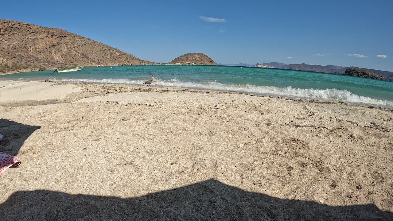 Baja California, Mexico beach with pale sand, a red towel edge entering from the left, turquoise surf, dry hills across the background, and a small person visible near the waves.