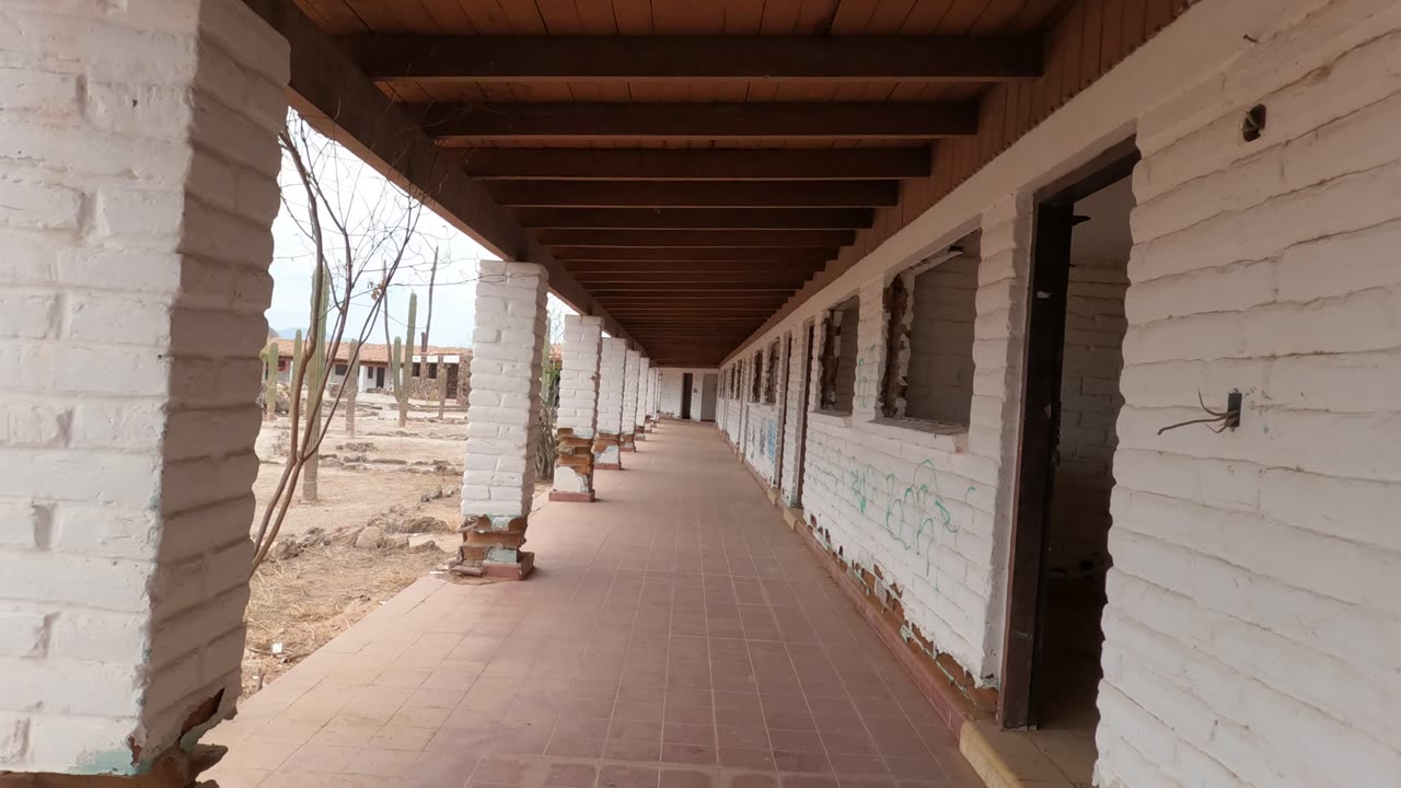 Opening view of a covered white brick walkway in Baja California, Mexico, with tiled floor, open doorways, dry ground, cacti, distant hills, and no visible people.