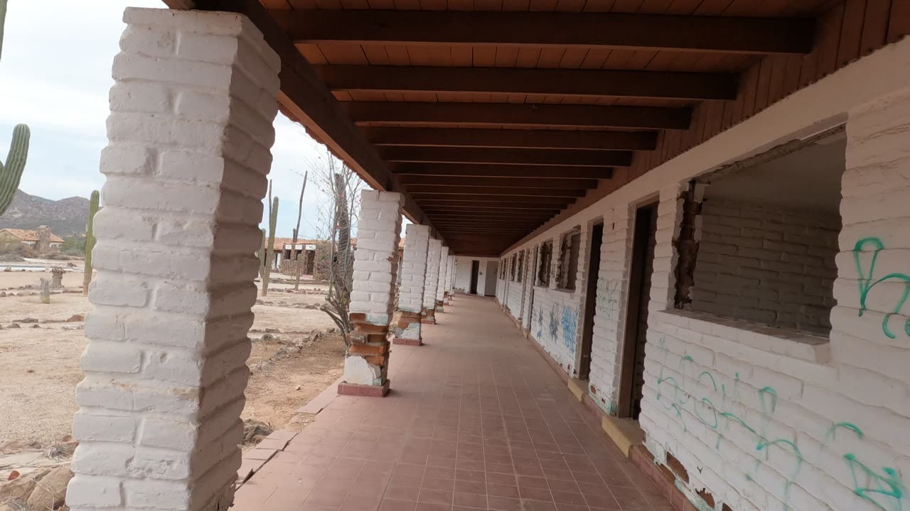 Start of a covered walkway in Baja California, Mexico, showing white brick columns, red floor tiles, graffiti, desert ground, cacti, distant mountains, and no visible people.