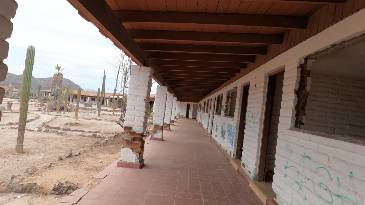 The walkway continues through Baja California, Mexico, with a white column near the foreground, dark window openings on the building, desert cactus plants outside, and no visible people.