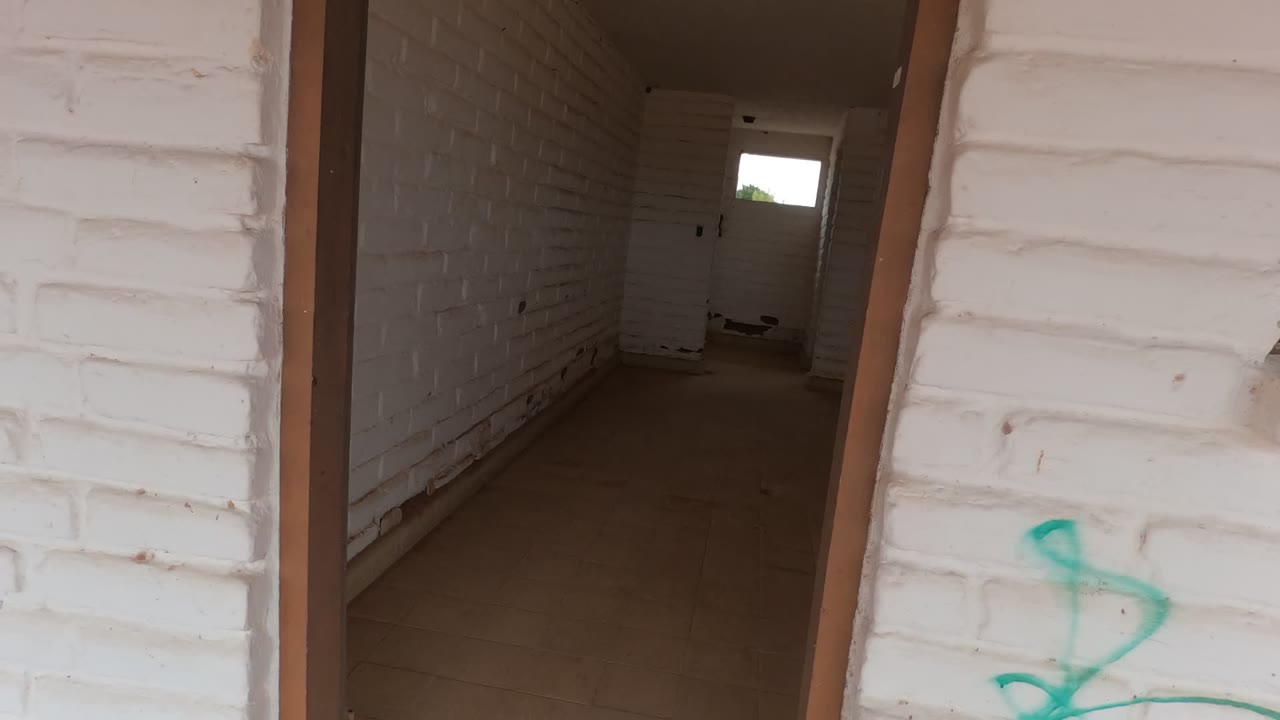 White brick doorway in a Baja California building opens into a dim empty room with a concrete floor, small rear window, and no visible people.