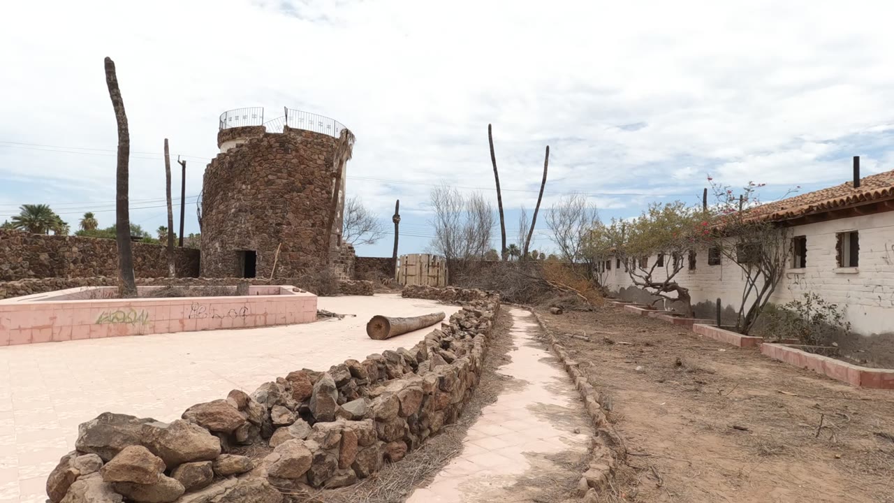 Wide view of a Baja California courtyard with a round stone tower, low rock wall, pink masonry planter, dry ground, sparse trees, and no visible people.