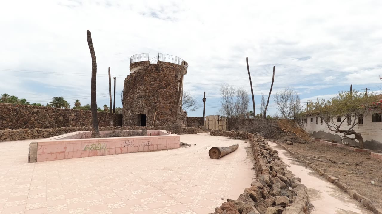 Wide Baja California courtyard frame with the stone tower left of center, a low white building with a tile roof, dry path, stone walls, and no visible people.
