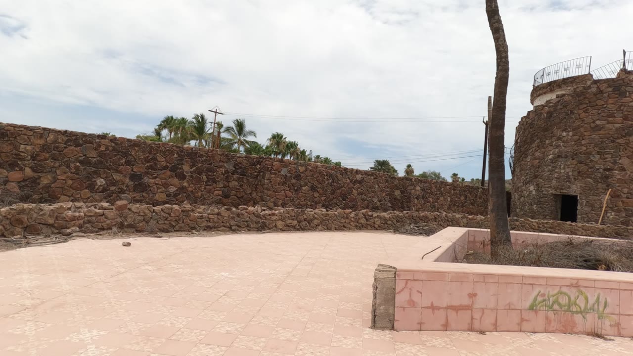 Baja California stone courtyard with pale pink paving, a palm trunk near center, rough enclosure walls, a circular tower at right, cloudy sky, and no visible people.