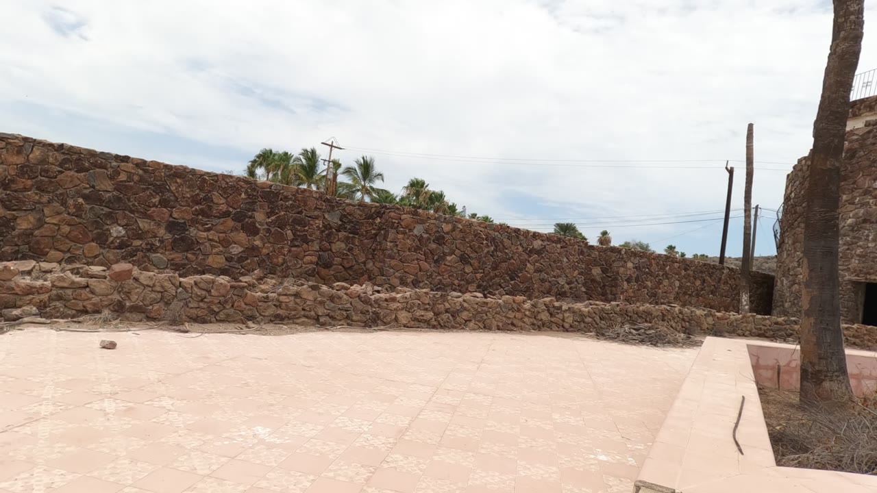 Baja California stone enclosure with a broad pale courtyard foreground, a circular tower with a dark doorway, palm trunks along the edges, utility lines, cloudy sky, and no visible people.