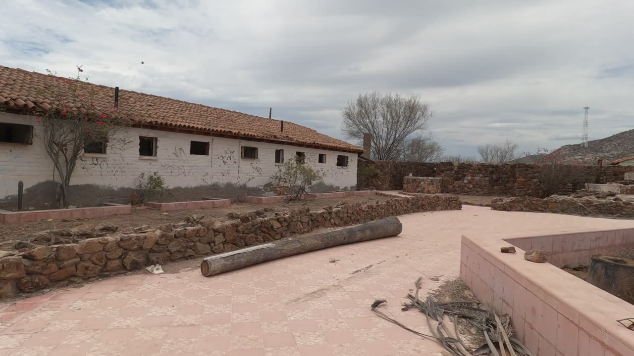 A Baja California courtyard shows a white building with a red tile roof, low stone walls, pale paving, a long log or pipe in the foreground, cloudy sky, and no visible people.