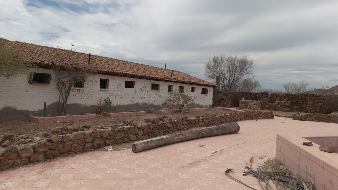 A white rural building in Baja California stretches across the frame behind stacked stone walls, with a bare tree, dry landscaping, cloudy sky, and no visible people.