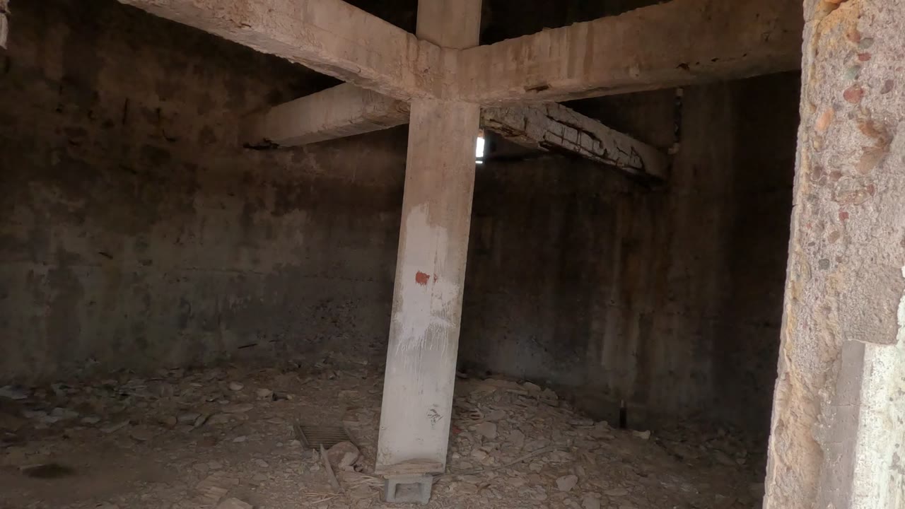 Dim room with rubble floor, concrete pillar, and beams.