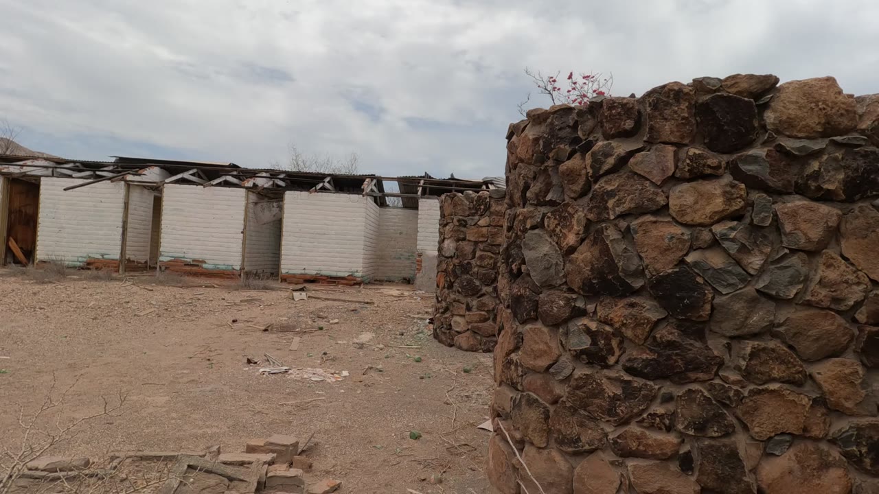 Baja California courtyard with a rough stone wall in the foreground, a damaged white brick building behind it, dry dirt ground, cloudy sky, and no visible people.