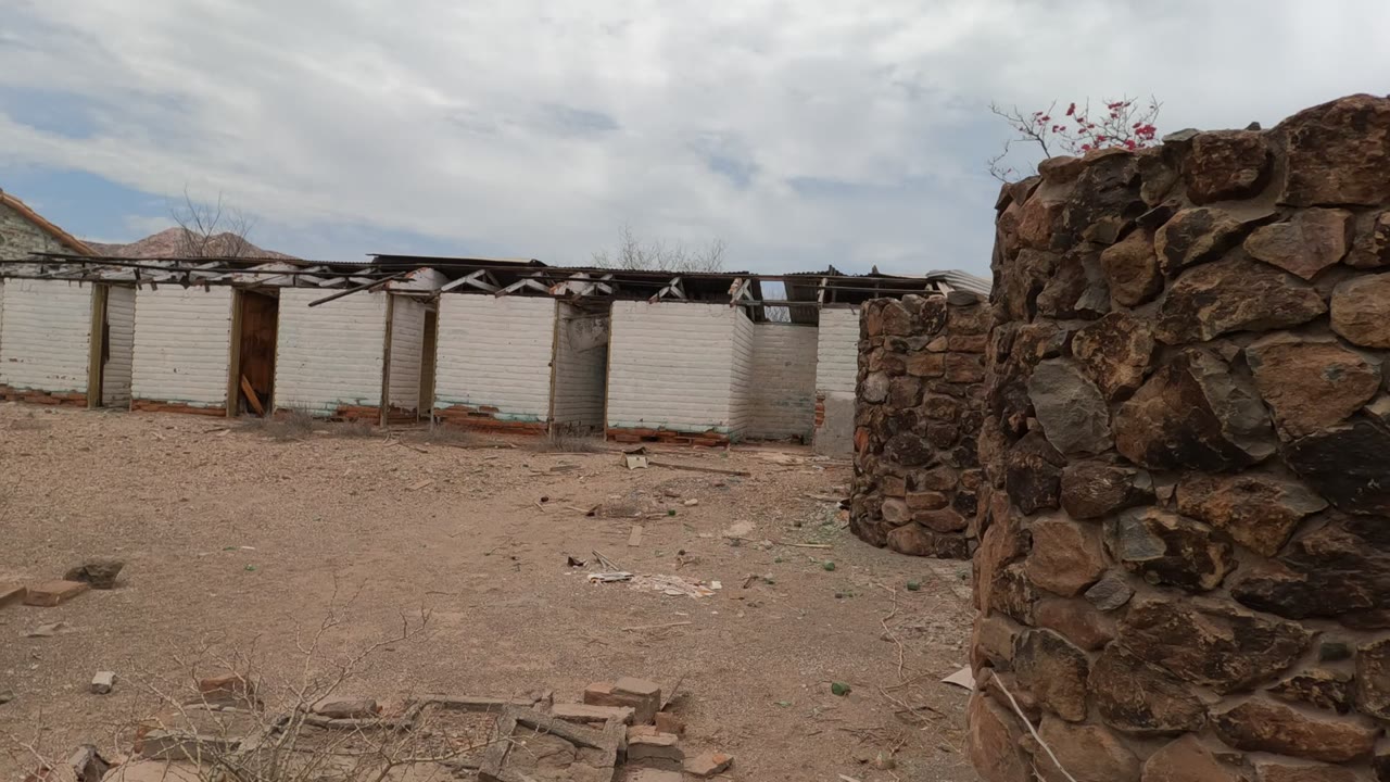 Baja California courtyard frame with the stone wall filling the right side, white brick structures receding across the background, dusty ground, broken materials, and no visible people.
