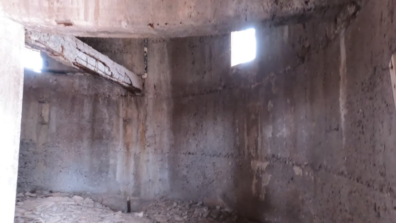 A wider upper-room view inside the abandoned Baja California concrete structure shows two bright wall openings, stained walls, ceiling edges, floor rubble, and no visible people.