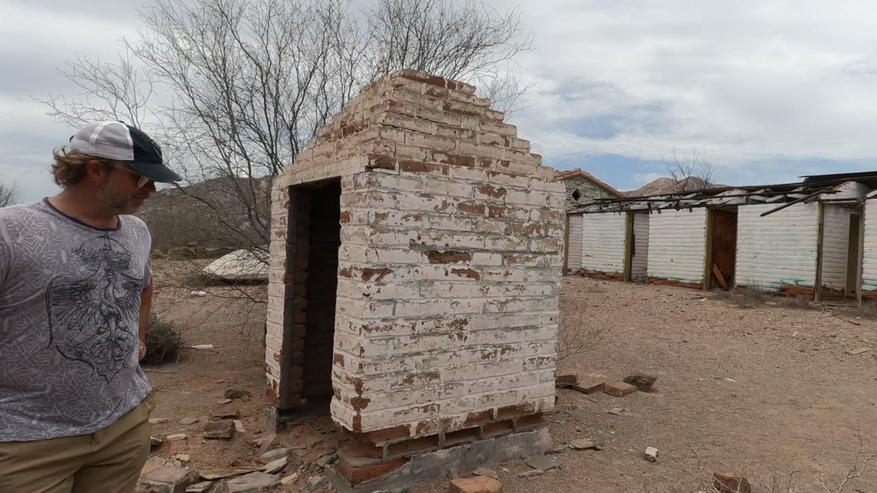 At 0:00, an adult man stands at the left edge beside a peeling white brick ruin in Baja California, Mexico, with dry ground and broken buildings beyond.