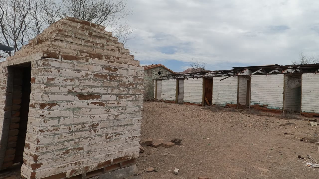 Peeling white brick wall and abandoned long building across a dirt yard.