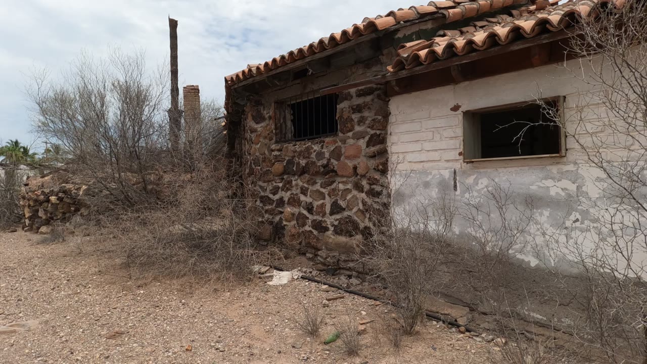 Weathered adobe building in Baja California, Mexico with a stone corner, white plaster wall, red tiled roof, dry brush, dirt ground, and no visible people.