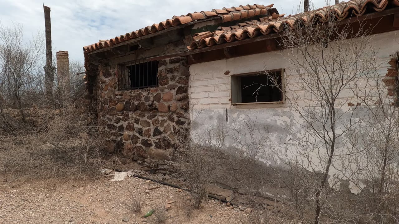 Side of abandoned plaster and stone building with barred window and leafless shrubs.
