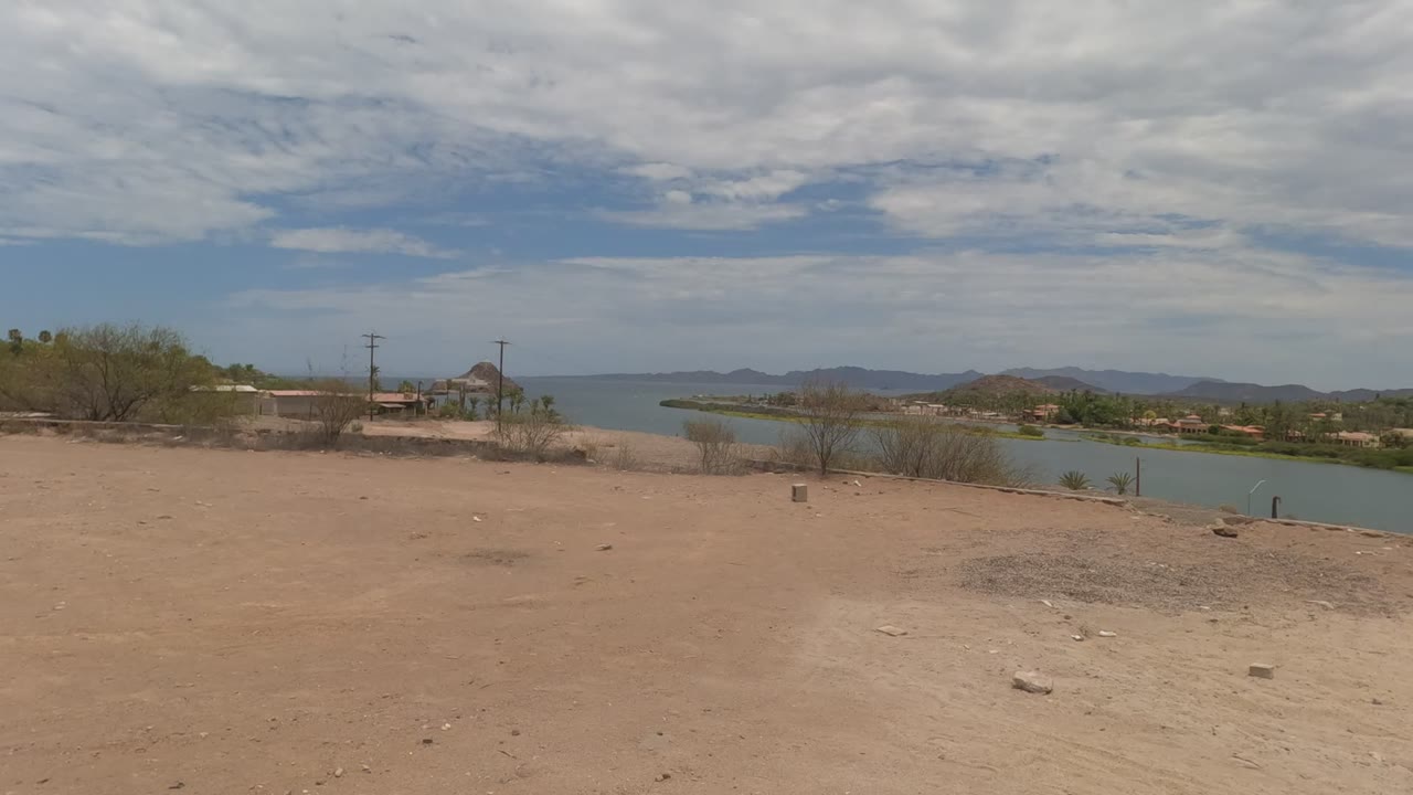 Wide Baja California desert lakeside view with sandy foreground, scattered rocks, water, distant mountains, layered clouds, and no visible people.