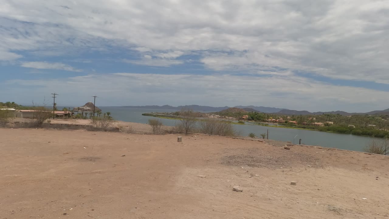 Arid lakeside ground with water, sparse trees, and low mountains.
