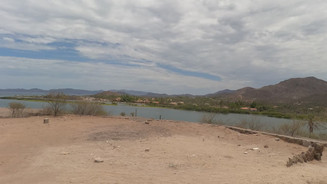 Baja California desert overlook with a broad sandy foreground, blue-green lake, low mountain range, scattered trees near the water, layered clouds, and no visible people.