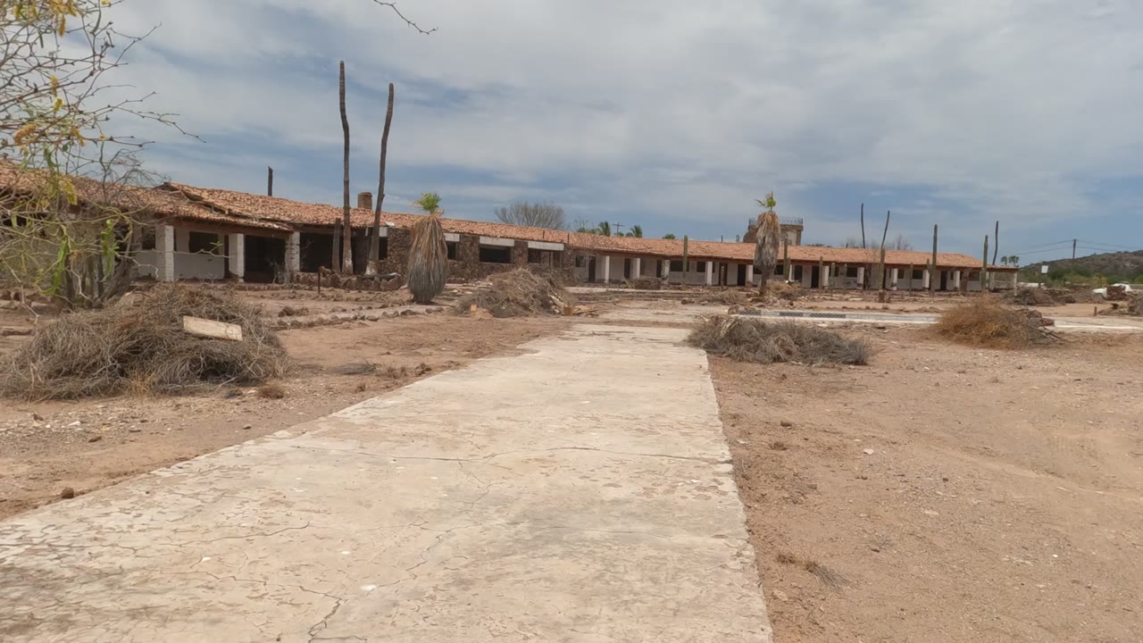 At the start of the Baja California, Mexico clip, a concrete path leads across sandy ground toward a long red-roofed abandoned building with dry brush and no visible people.