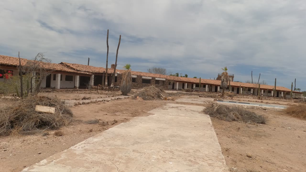 The Baja California, Mexico frame shows sandy ground and brush in the foreground, the concrete path angling toward the long abandoned building, tall cacti, and no visible people.