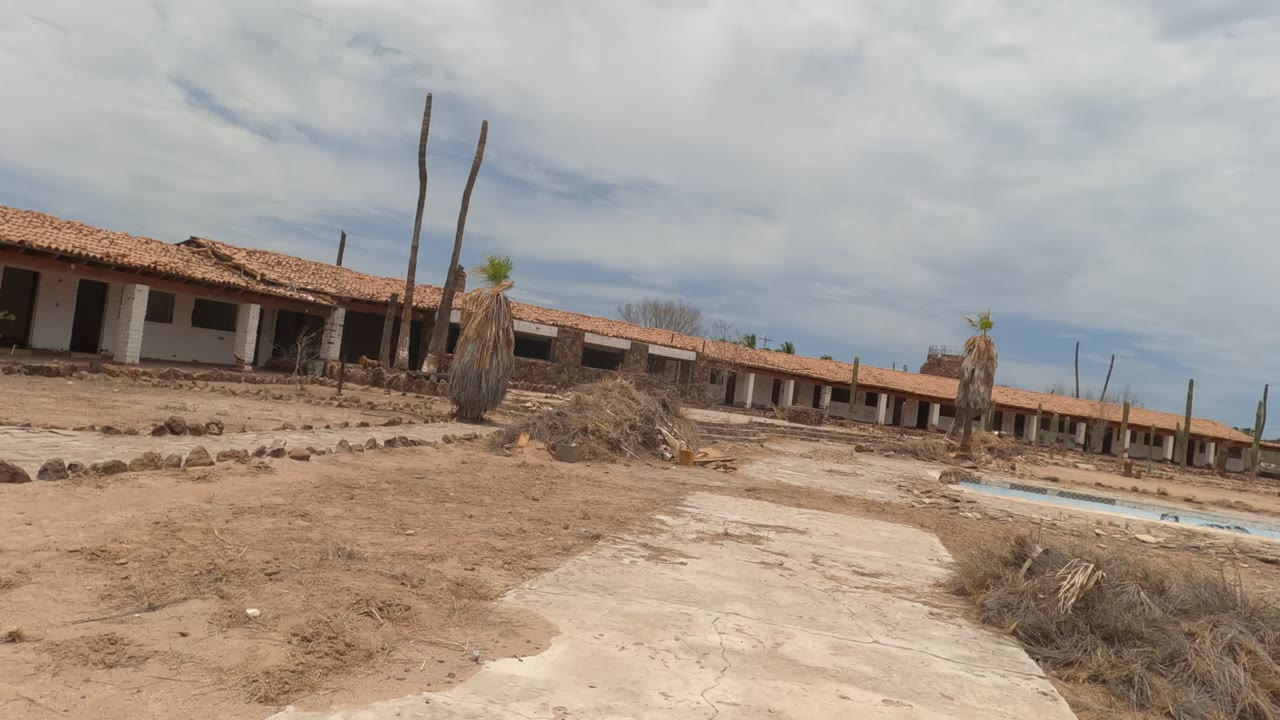 Wide view in Baja California, Mexico of an abandoned red-tile-roof motel beside dry sandy terrain, a drained pool, sparse palms, and no visible people.