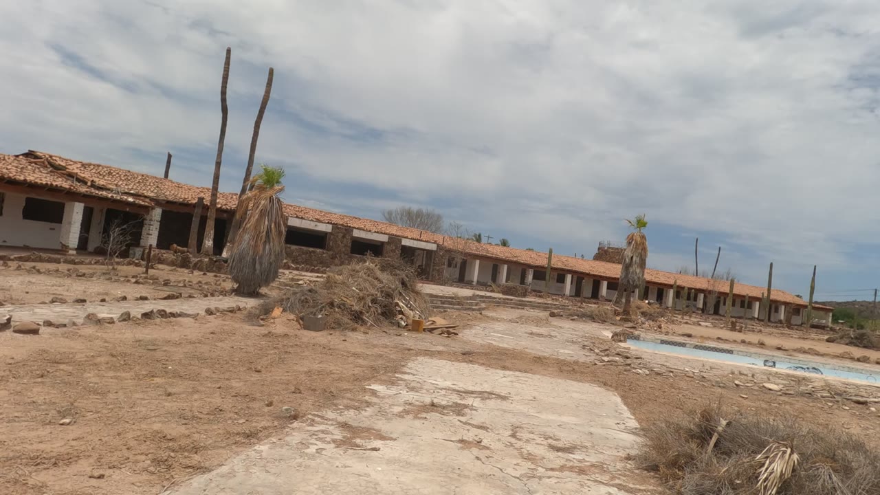 Wide Baja California frame of a dilapidated motel row, sandy open ground, a blue-edged drained pool, sparse palms, and an overcast sky with no visible people.