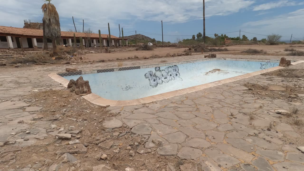 Wide view of an abandoned dry swimming pool in Baja California, Mexico, with pale blue walls, graffiti, rocks on cracked decking, low buildings in the background, and no visible people.