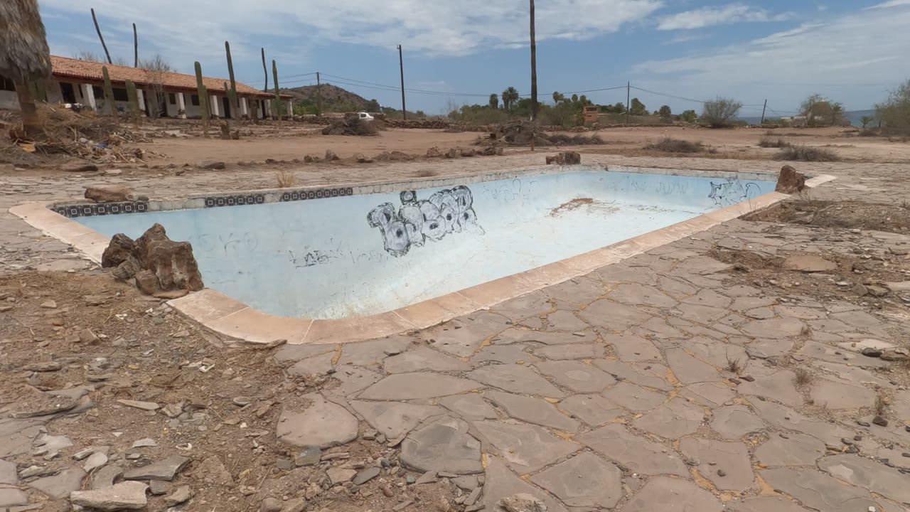 Dry blue pool and rock foreground with abandoned buildings across a dirt lot.