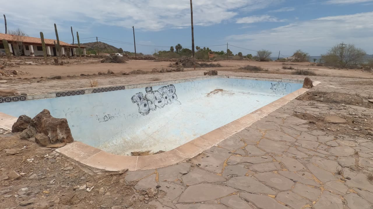 Slightly shifted view of the abandoned pool in Baja California, Mexico, with graffiti on the inner wall, cracked deck stones in front, low buildings to the left, and no visible people.