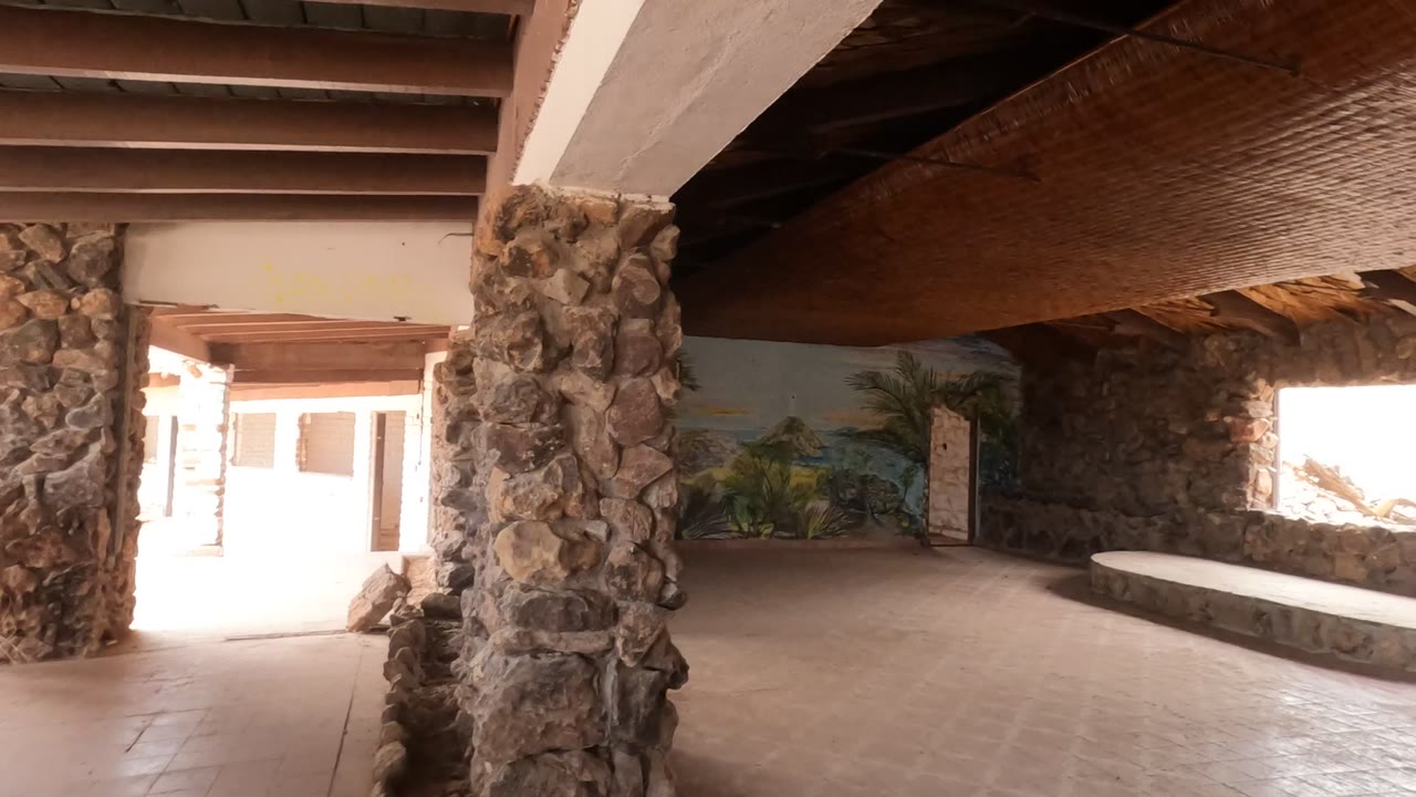 Opening frame of a Baja California, Mexico interior with rough stone pillars in the foreground, tiled floor, a bright doorway at left, thatched ceiling above, and no visible people.