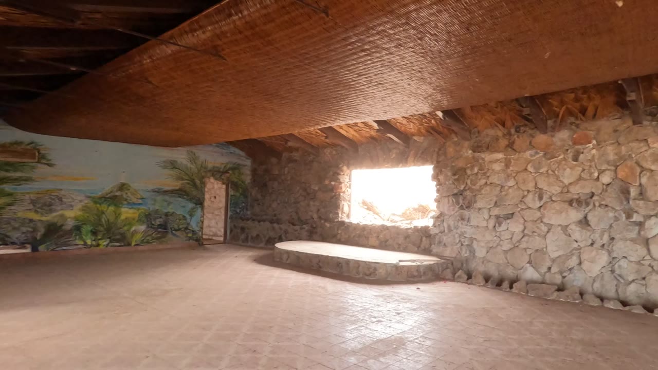 Mid-pan view inside a Baja California, Mexico stone room with a low bench beneath a bright window, a mural along the back wall, tiled floor, overhead thatch, and no visible people.