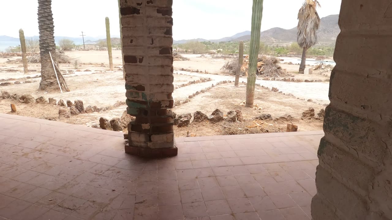 Shaded patio in Baja California, Mexico with brick columns, reddish floor tile, sandy garden beds, tall cacti, distant mountains, and no visible people.