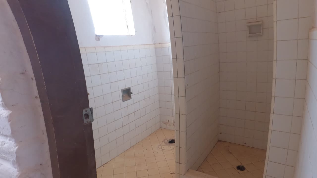 White tiled bathroom stalls in Mulege, Mexico, with beige floor tiles, a dark door edge, and bright daylight from a high window, no visible people.
