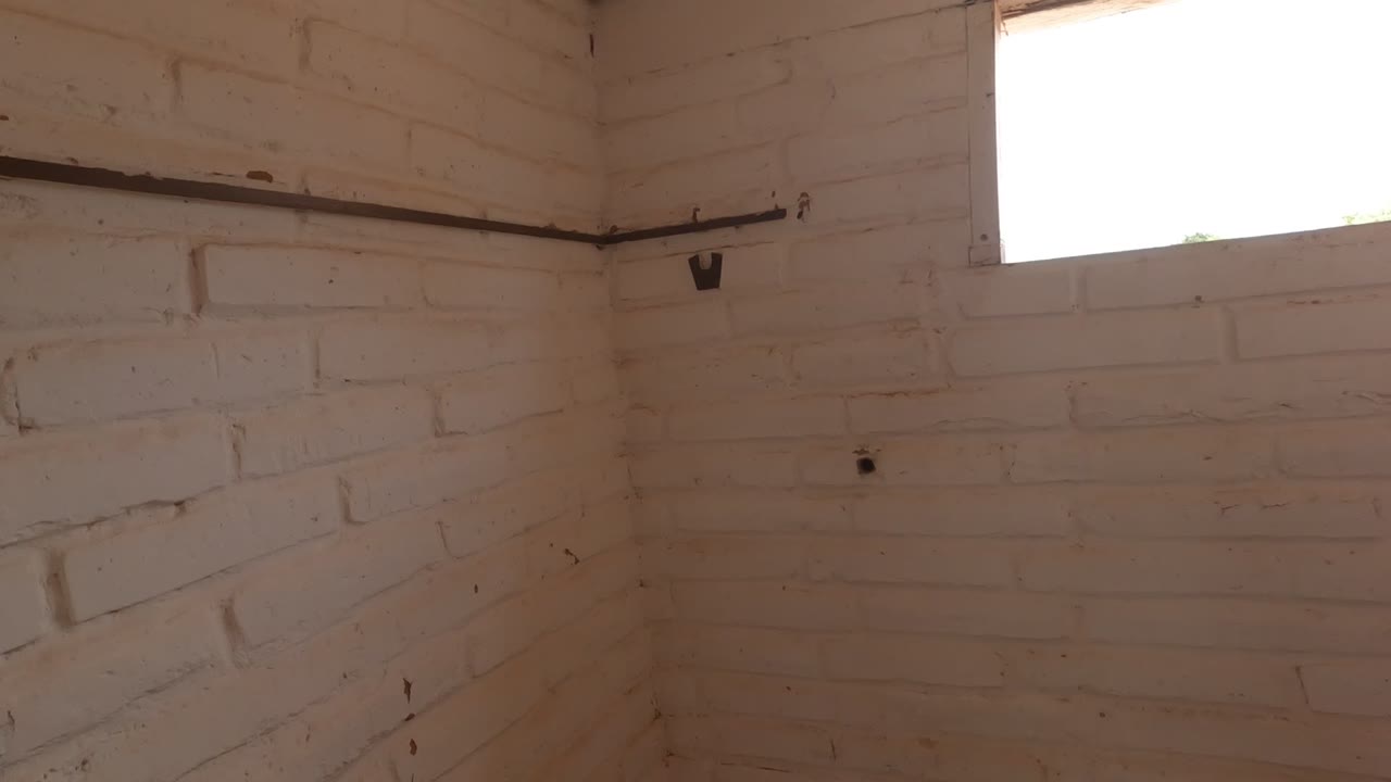 Corner view of the Mulege bathroom showing white-painted brick, a high window, dark wall rail, small holes, and worn paint near the floor, no visible people.