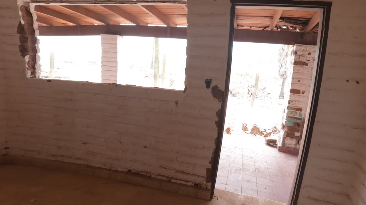 Unfinished white brick interior in Mulege with a dark doorway frame, bright outdoor patio light, rectangular window opening, and no visible people.