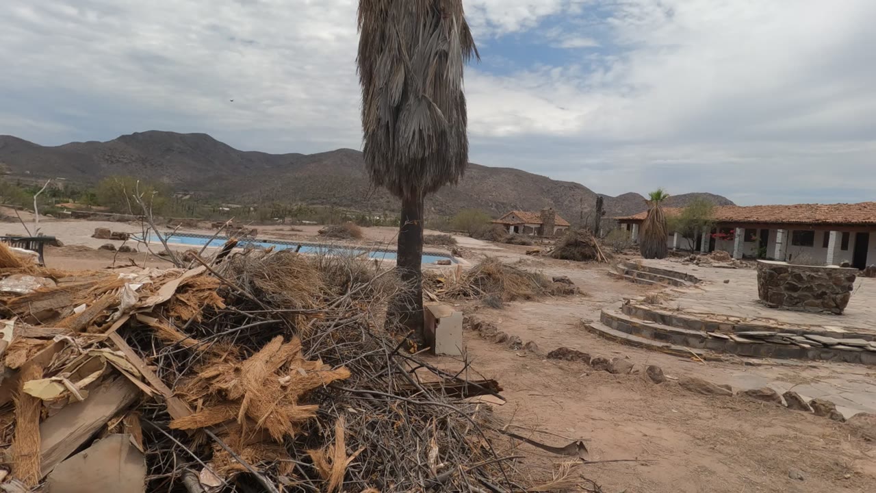 Opening view in Baja California, Mexico shows a debris pile and palm trunk in the foreground with a pool edge, low desert buildings, distant mountains, cloudy sky, and no visible people.