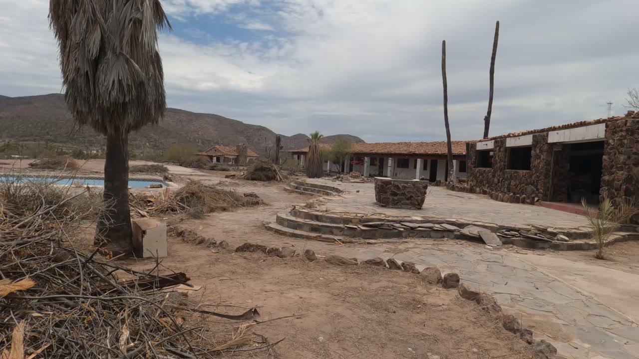 The pan reaches a wider Baja California courtyard view with a palm trunk near center, stone-bordered paths, dry brush, low stucco and stone buildings, mountains, cloud cover, and no visible people.