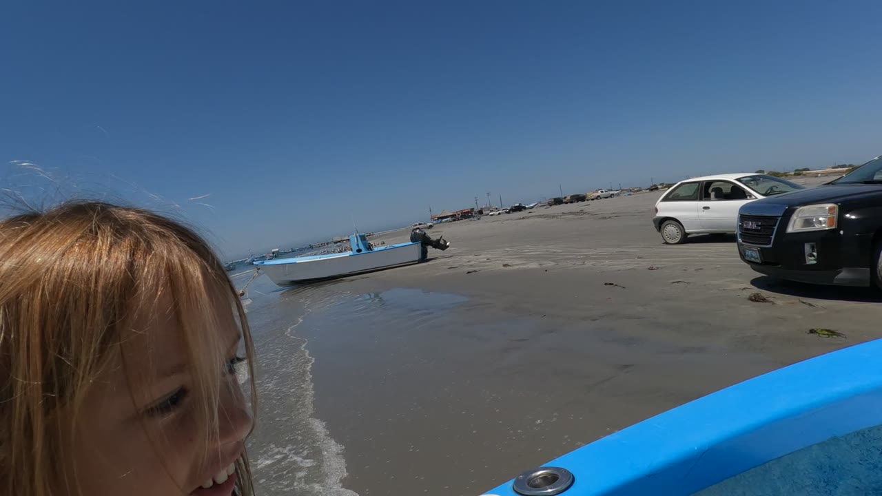 A young child appears at the edge of a blue boat in Baja California, Mexico, with wet beach sand, parked cars, and another small boat in the background.