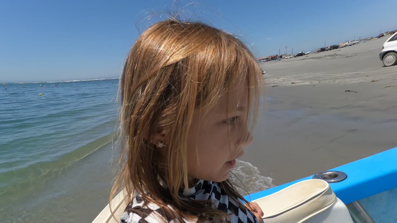 The child is shown in side profile on the blue boat in Baja California, Mexico, with rippled ocean water on the left and a sandy beach on the right.