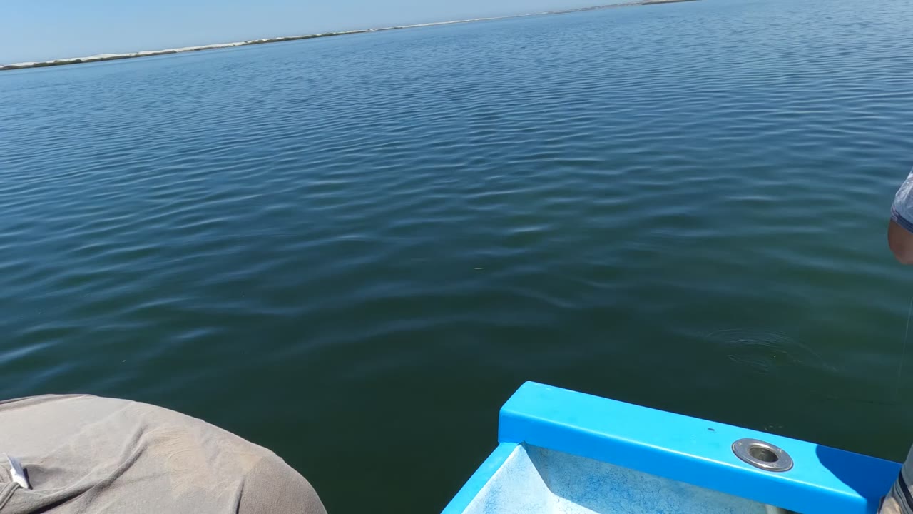 Opening frame in Baja California, Mexico showing rippled blue water, a low distant shoreline, and the bright blue edge of a small boat with only part of a seated person visible at the lower left.