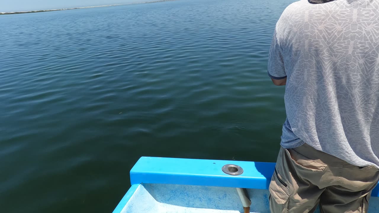The man remains near the right side of the Baja California boat frame, with the bright blue rail below him and calm open water stretching to a thin shoreline.