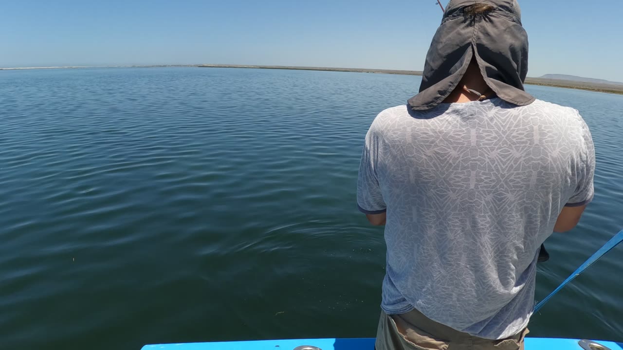 Hooded person holds a fishing rod over rippled water from a blue boat.