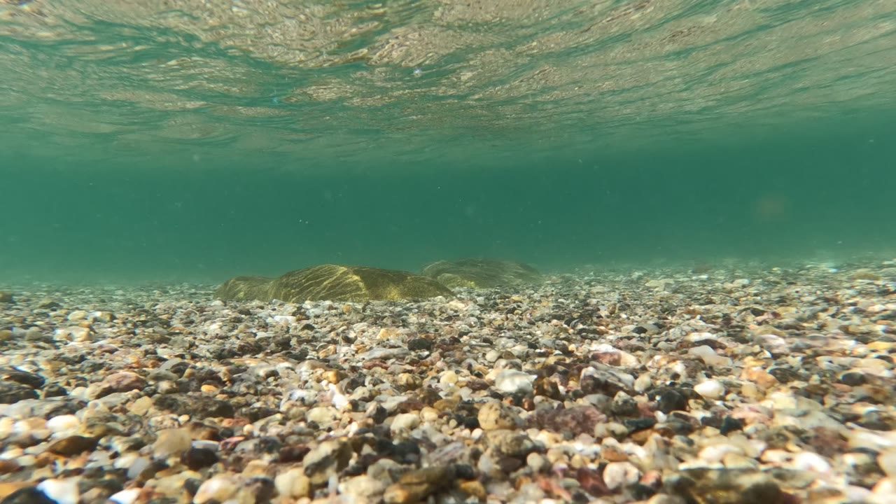 Split-level moment in Baja California, Mexico, where water covers much of the lens, blurring the pebbly foreground while the ocean horizon and distant land remain partly visible, with no visible people.