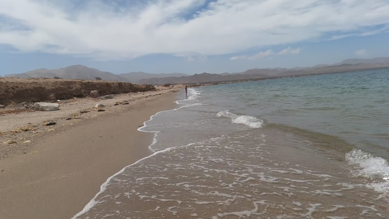 Baja California, Mexico beach view with white foam tracing the wet sand, turquoise water to the right, low rocky shore to the left, and one distant person near the shoreline.