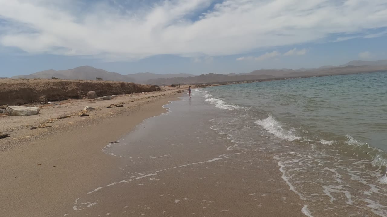 Baja California, Mexico beach frame with wave foam pushing farther along the right edge, smooth wet sand in the center, a distant person near the waterline, and hazy hills beyond the shore.