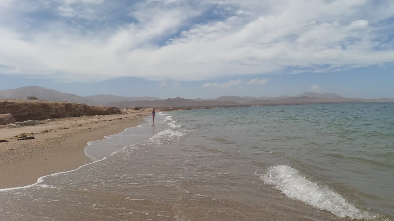 Baja California, Mexico beach at 0:00 with a low view of wet sand, gentle surf, one distant person near the waterline, turquoise ocean, clouds, and hazy mountains.