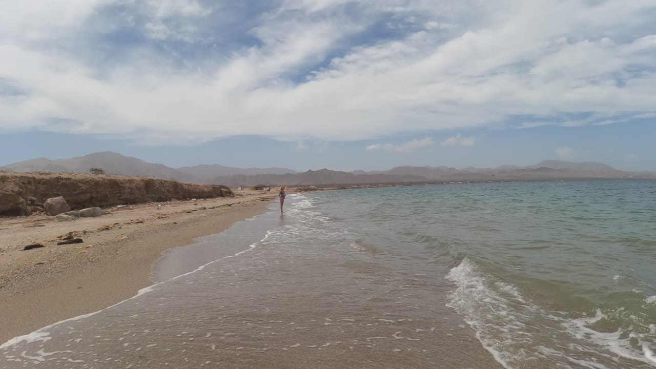 Small wave reaches the shore beside a distant person under a cloudy sky.